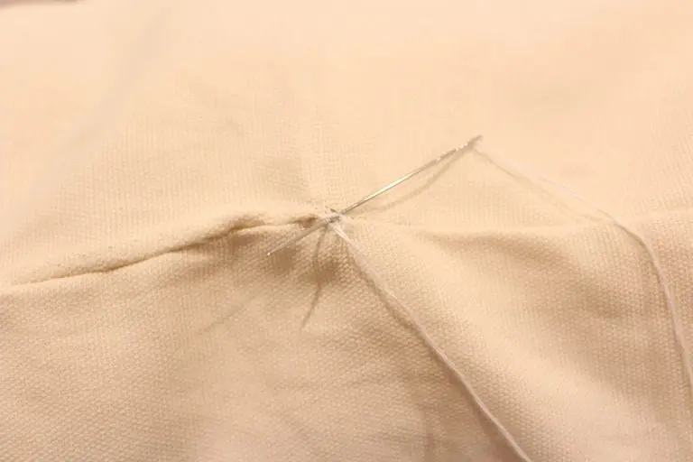Close-up of hand stitching a seam with a needle and white thread while making a DIY throw pillow.