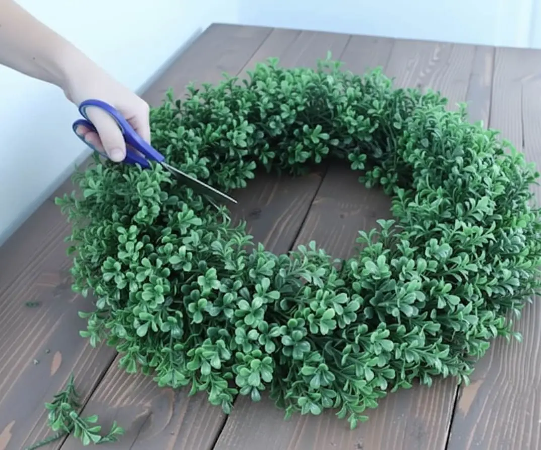 Hands using blue scissors to trim and shape a newly made DIY boxwood wreath, ensuring an even and full appearance for the faux greenery.