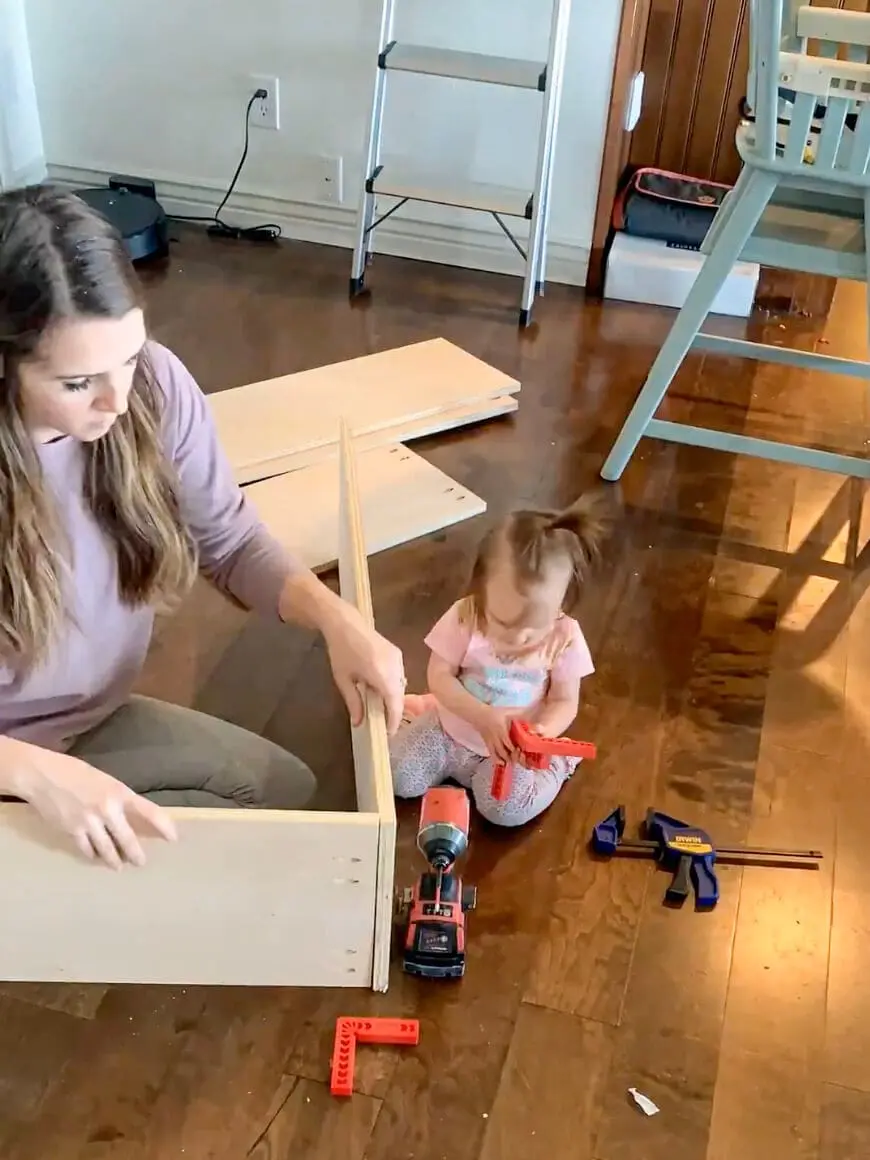 Woman assembling a wooden cabinet box on a hardwood floor, preparing for built-in office storage.