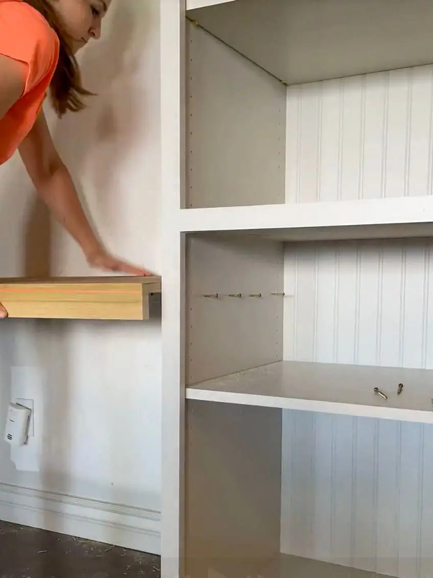Woman securing a wooden floating desk between built-in cabinets with a power drill.