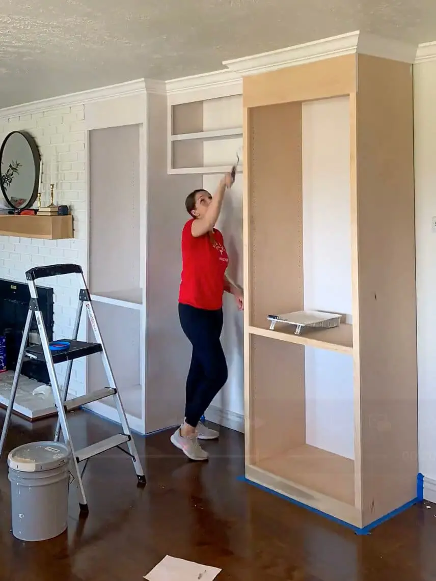 Woman painting a tall built-in cabinet with a roller while standing on the floor.