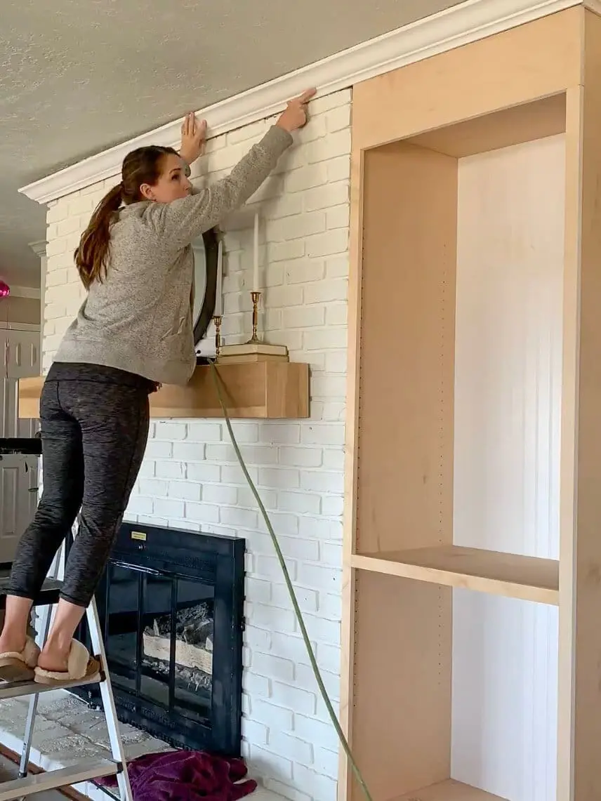 Woman installing crown molding on a built-in bookshelf next to a fireplace using a nail gun.