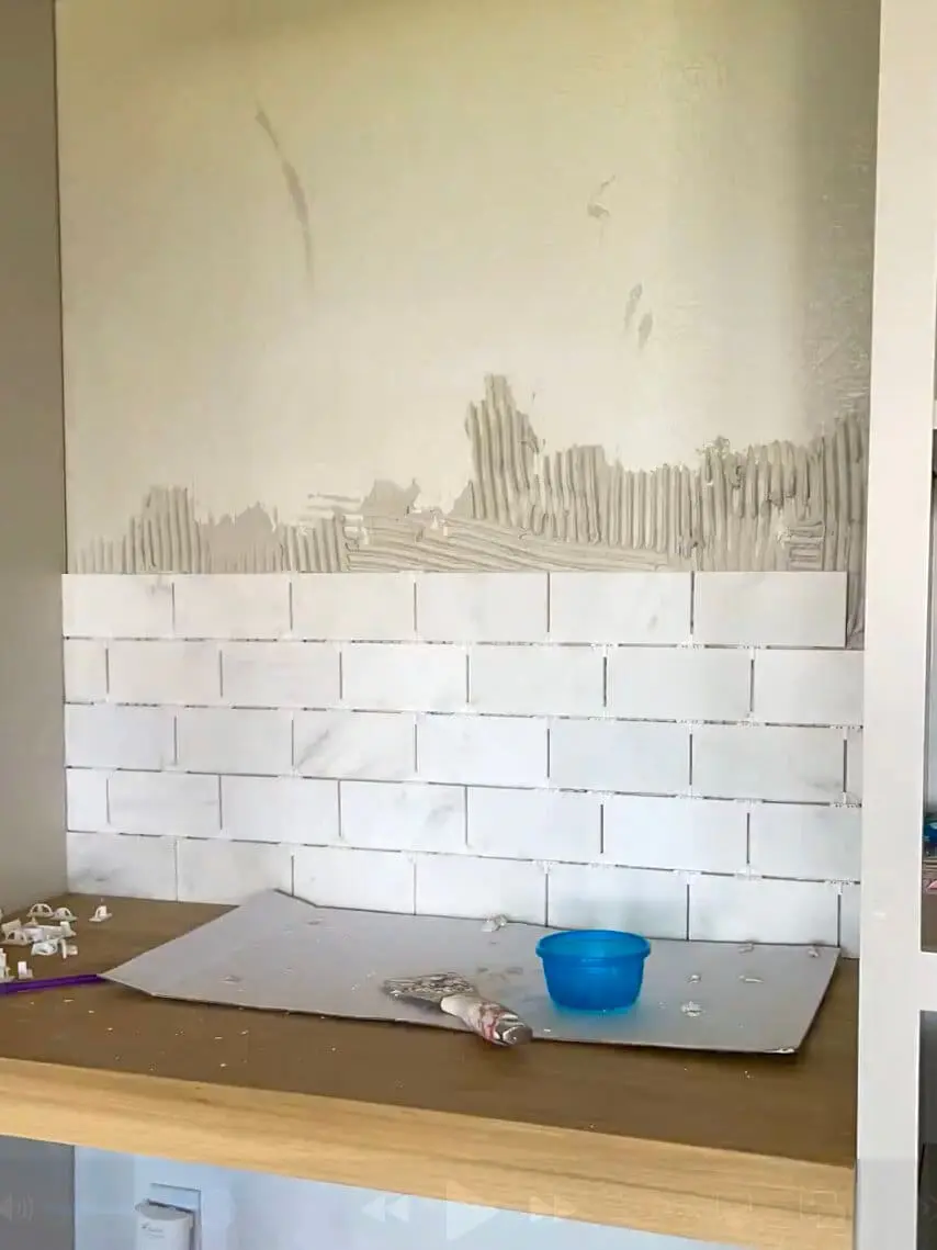 Partially installed marble subway tile backsplash above a wooden desk