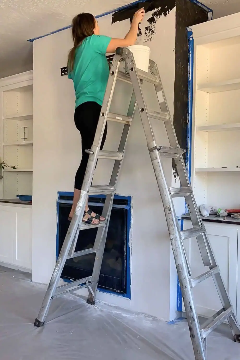 Woman on ladder applying black plaster finish to top of fireplace wall, bucket of clay on ladder shelf, fireplace insert taped off with blue tape