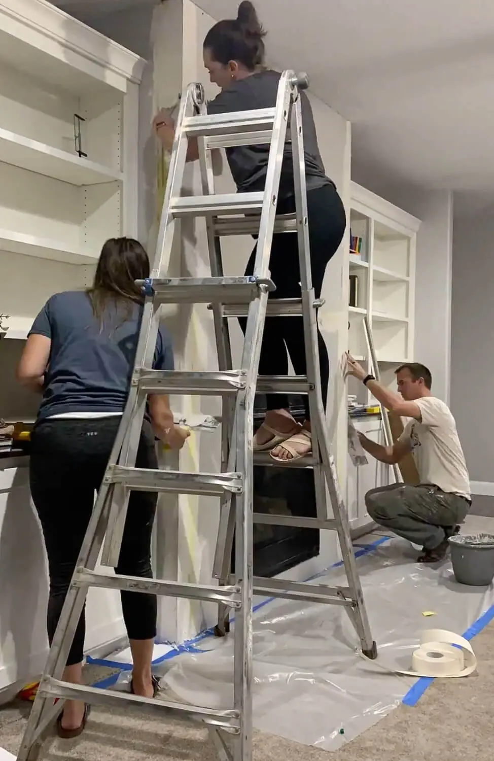 Three people prepping fireplace for renovation - one on ladder, two below taping and positioning, plastic drop cloth on floor