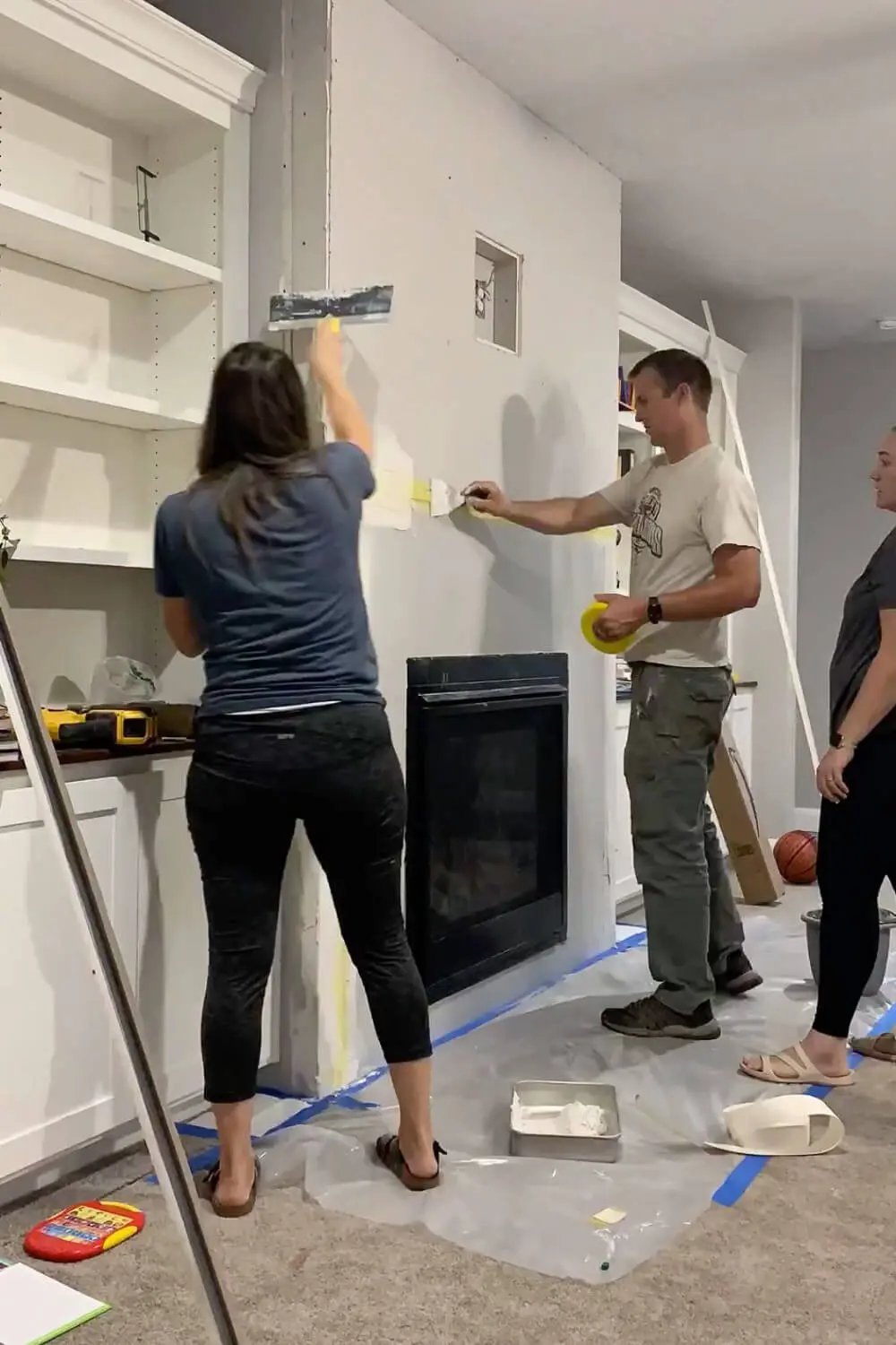 Multiple people applying joint compound to fireplace drywall - woman with taping knife spreading mud while man holds painters tape