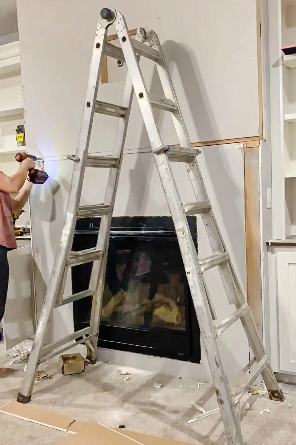 Person using cordless drill to secure large sheet of drywall to fireplace wall framing, ladder positioned in front of fireplace