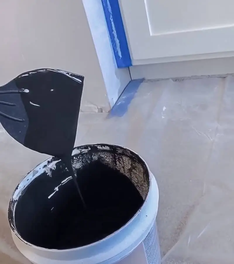 Plastic putty knife being pulled from bucket showing thin black plaster consistency, plastic drop cloth and blue tape in background