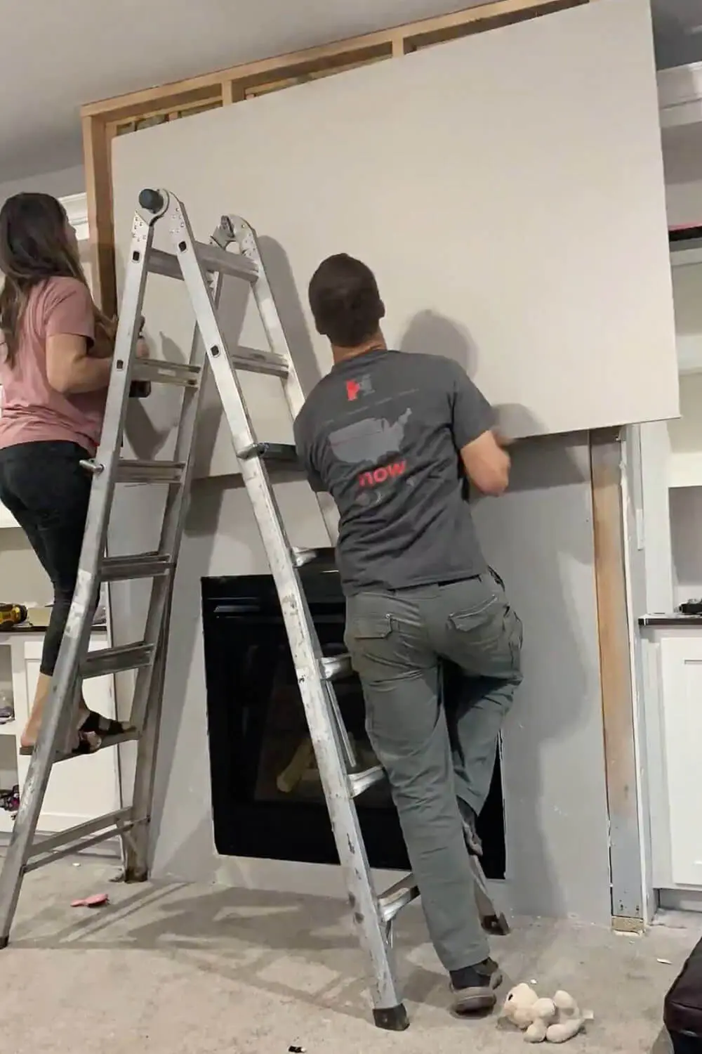 Two people lifting large drywall sheet onto fireplace wall - woman on ladder holding top edge, man positioning from below