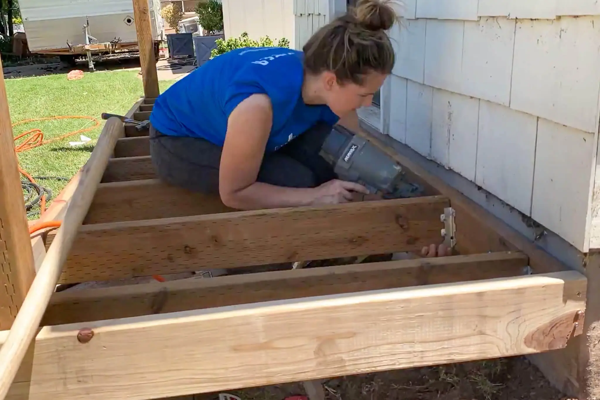 Woman crouched inside the joist frame of a partially built front porch, using a nail gun to secure a joist hanger to the ledger board against the house.