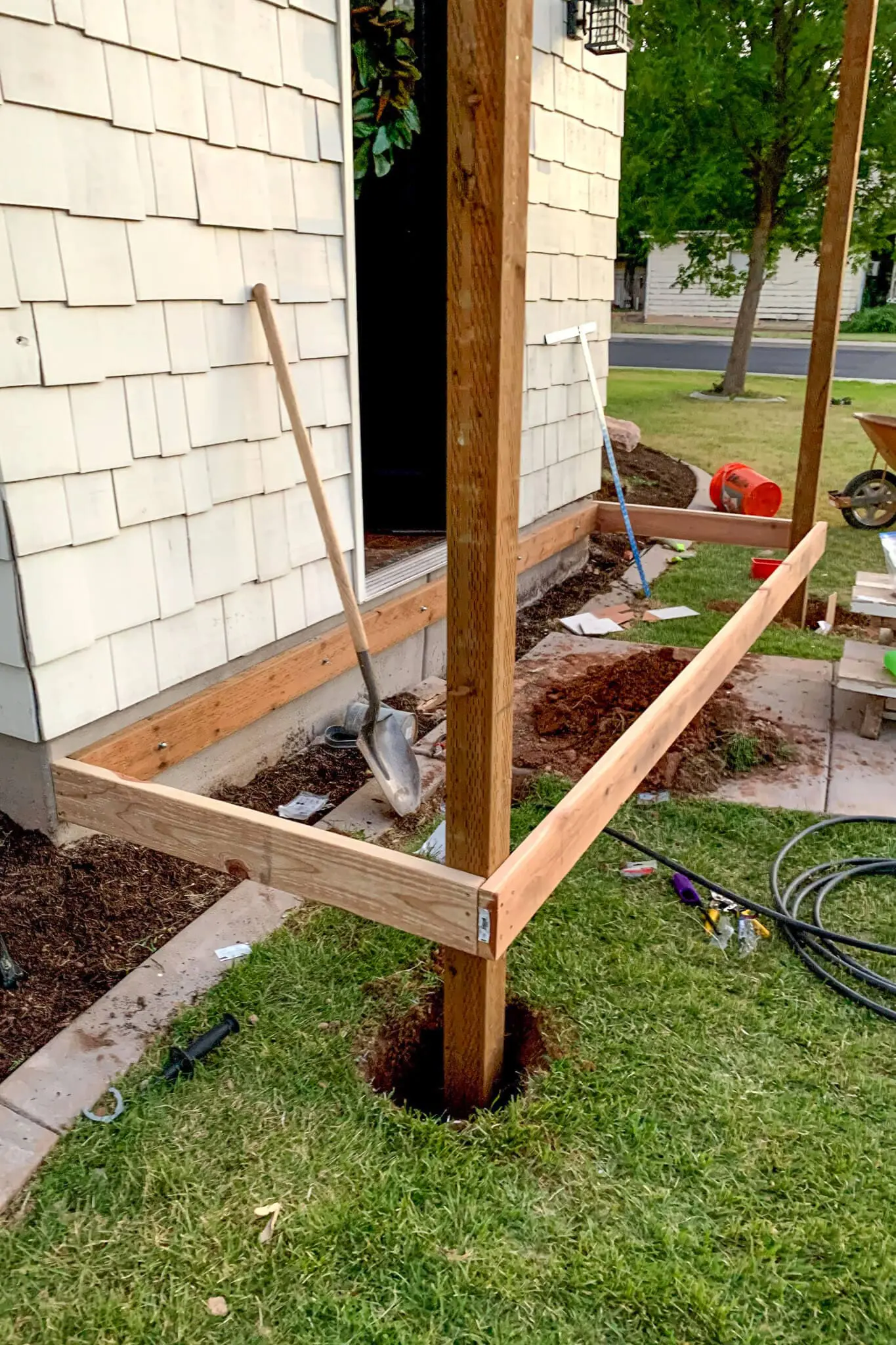 Framed DIY front porch in progress showing vertical 4x4 posts set in concrete holes and rim joists attached, with tools and a shovel nearby