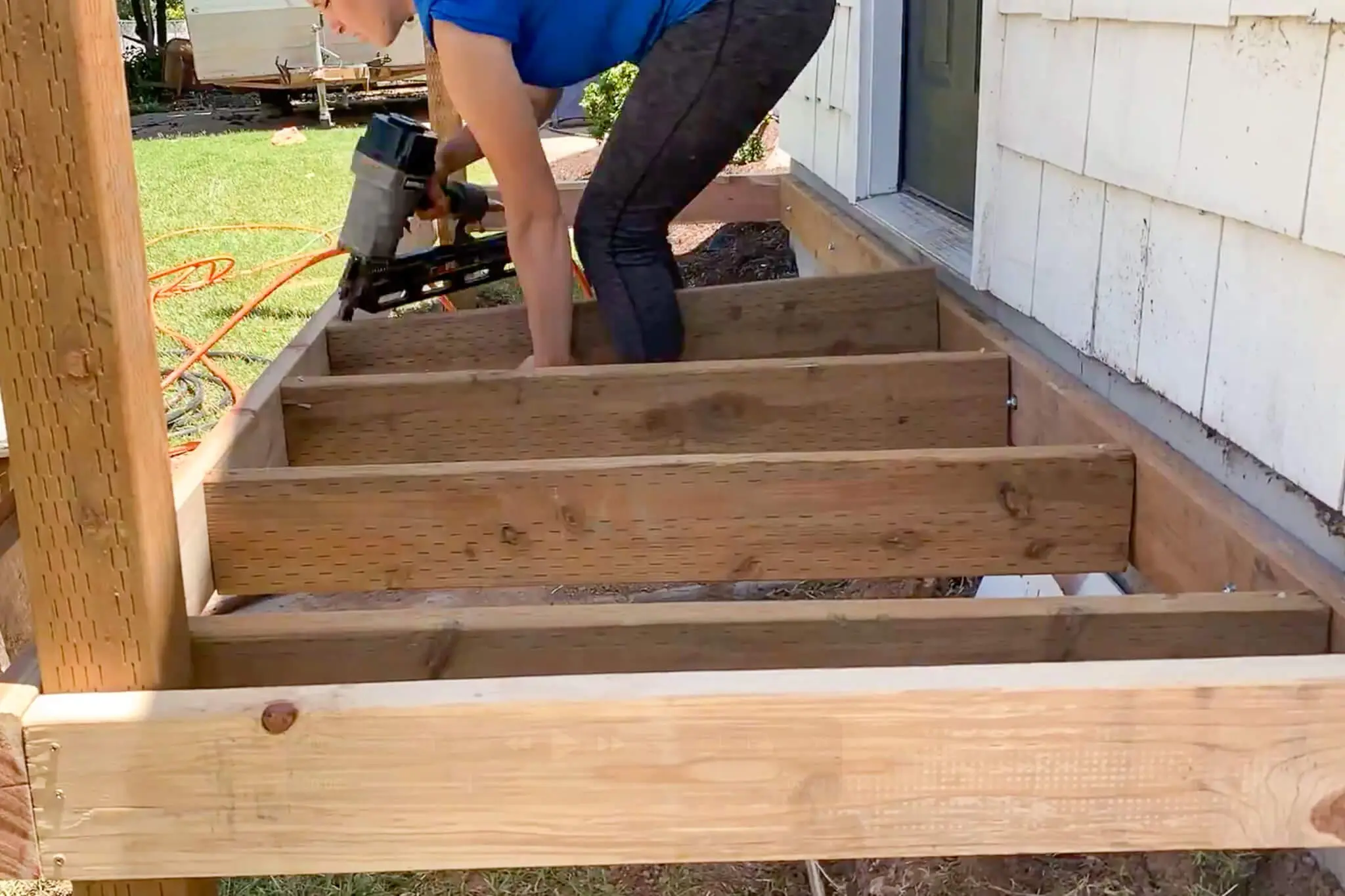 Woman using a nail gun to secure joists on a partially built wooden front porch deck, with unfinished frame and visible ledger board along the house.