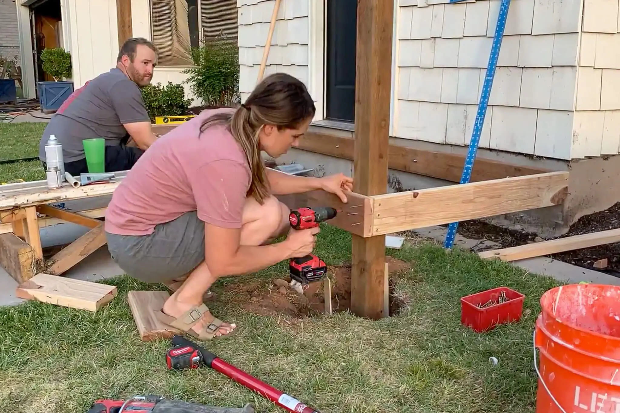 Framing a DIY front porch with wood posts and rim joists in place, showing shovel resting inside post hole and tools scattered on the grass during construction.