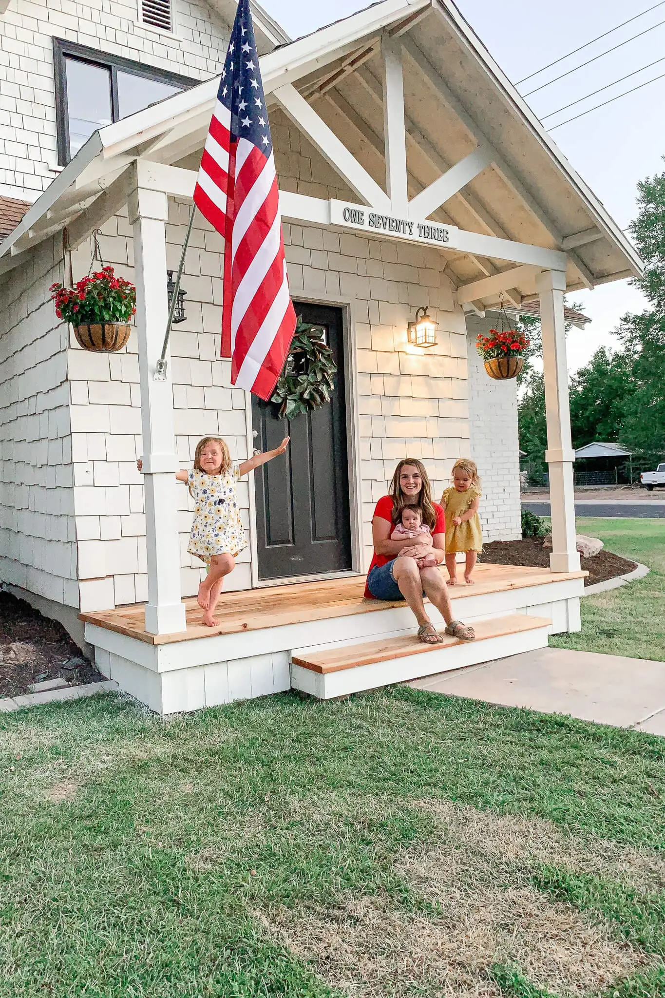 Family sitting on newly built white front porch with black door and American flag, showcasing a charming DIY home exterior upgrade.