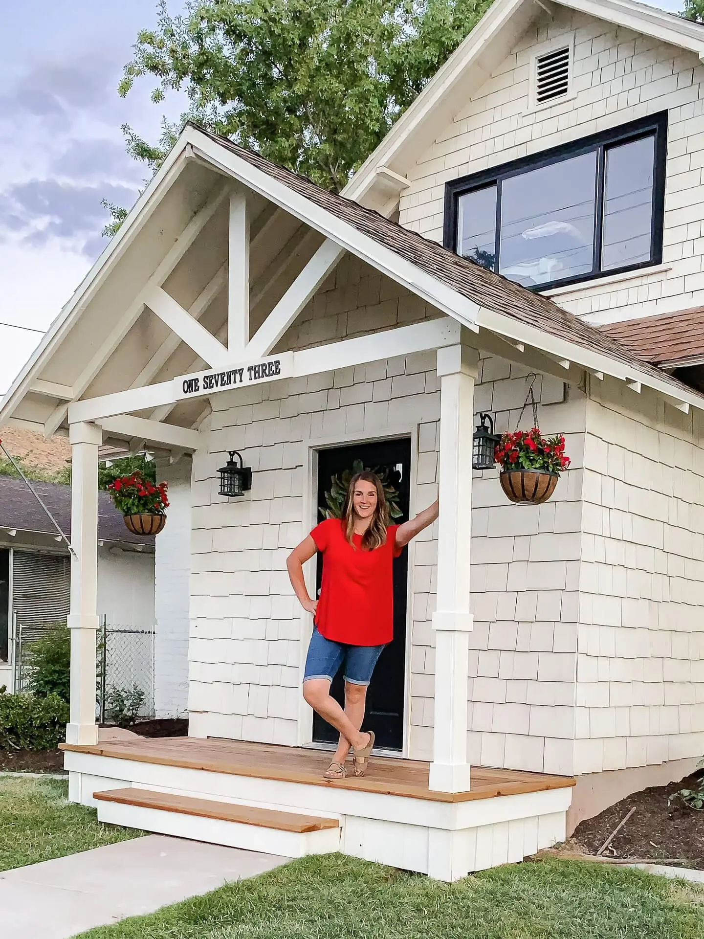 Woman standing on a finished DIY small front porch with white trim, wood decking, and hanging flower baskets, in front of a cream shingle-style house.