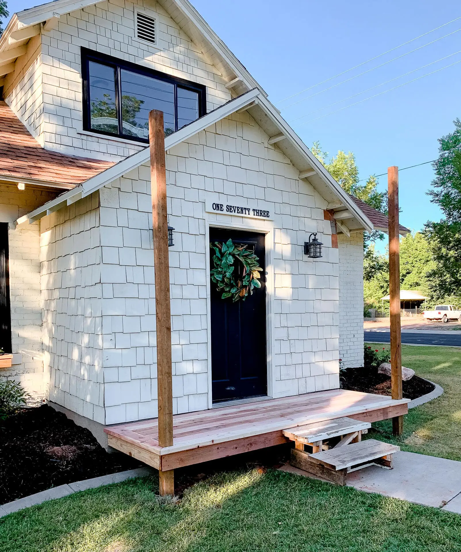 DIY small front porch with completed deck boards and exposed vertical posts, shown attached to a cream shingle-style house with a dark front door and temporary wooden steps.