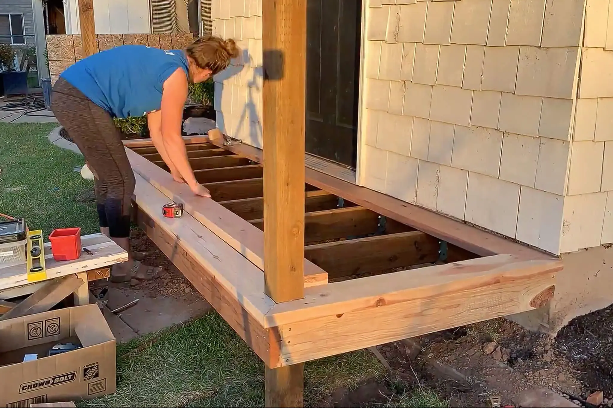 Woman placing deck boards during a DIY small front porch build, aligning boards across exposed joists with tools and materials nearby.