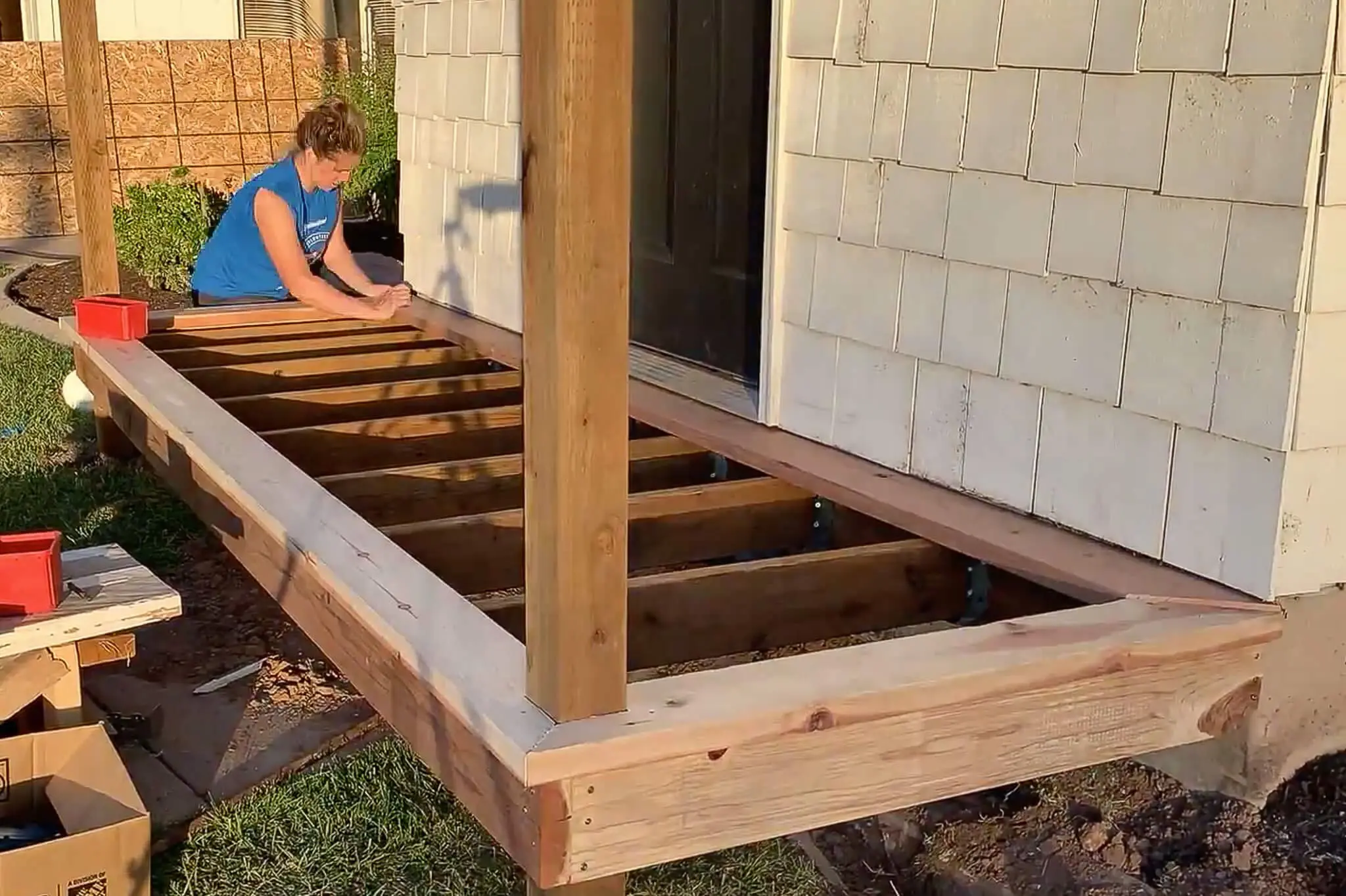 Woman marking a board during the trim installation phase of a DIY small front porch build, with the porch frame and joists fully exposed.