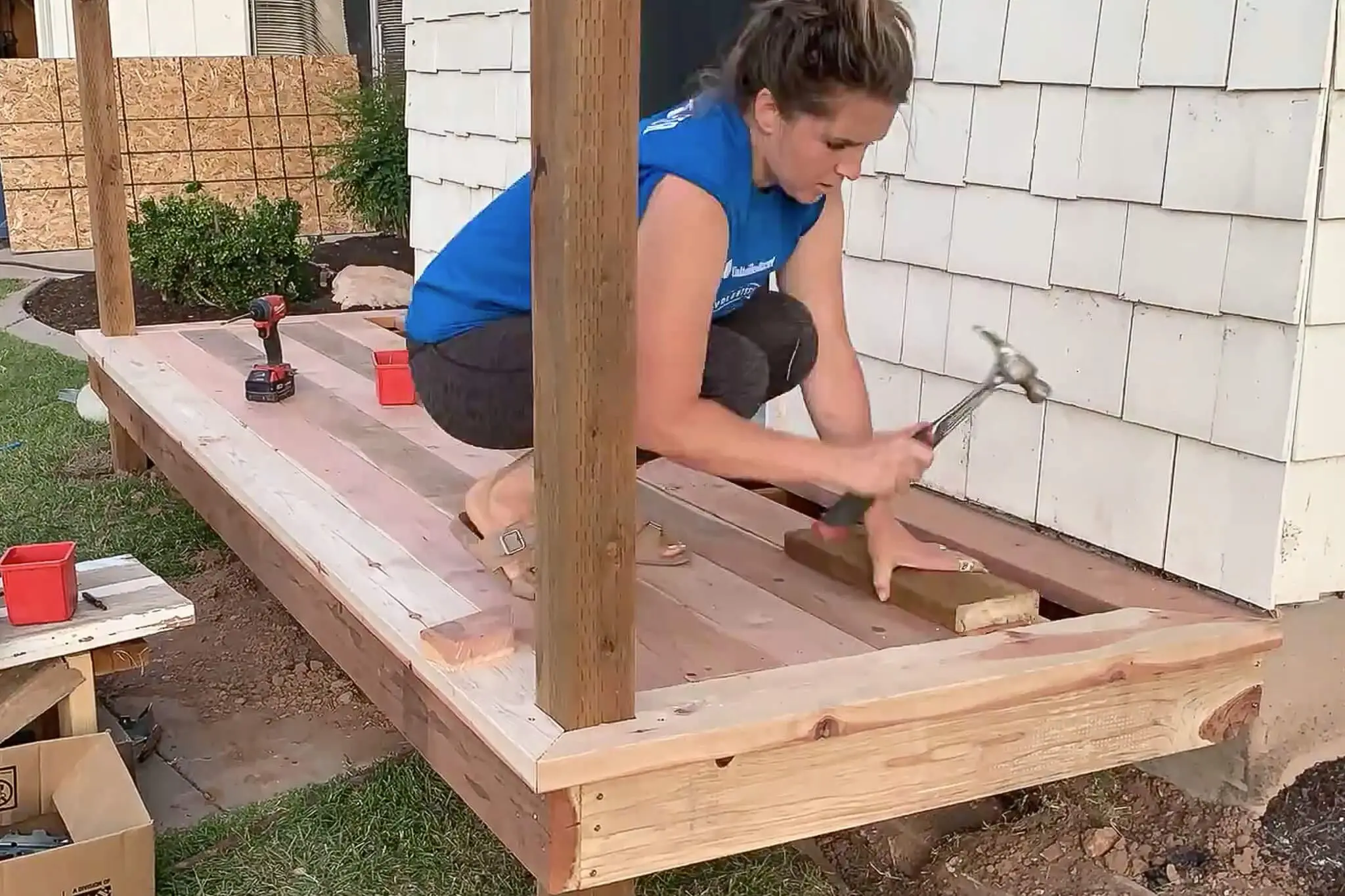 Woman using a hammer and scrap wood to tap a deck board into place during a DIY small front porch project, with nearly completed decking visible.