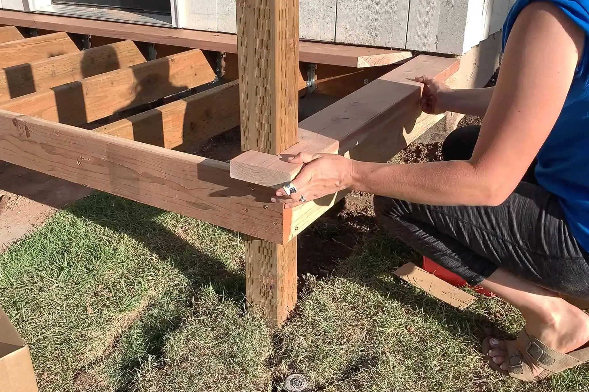 Woman positioning a notched trim board around a porch post during the construction of a small front porch, with exposed joists and frame in progress.
