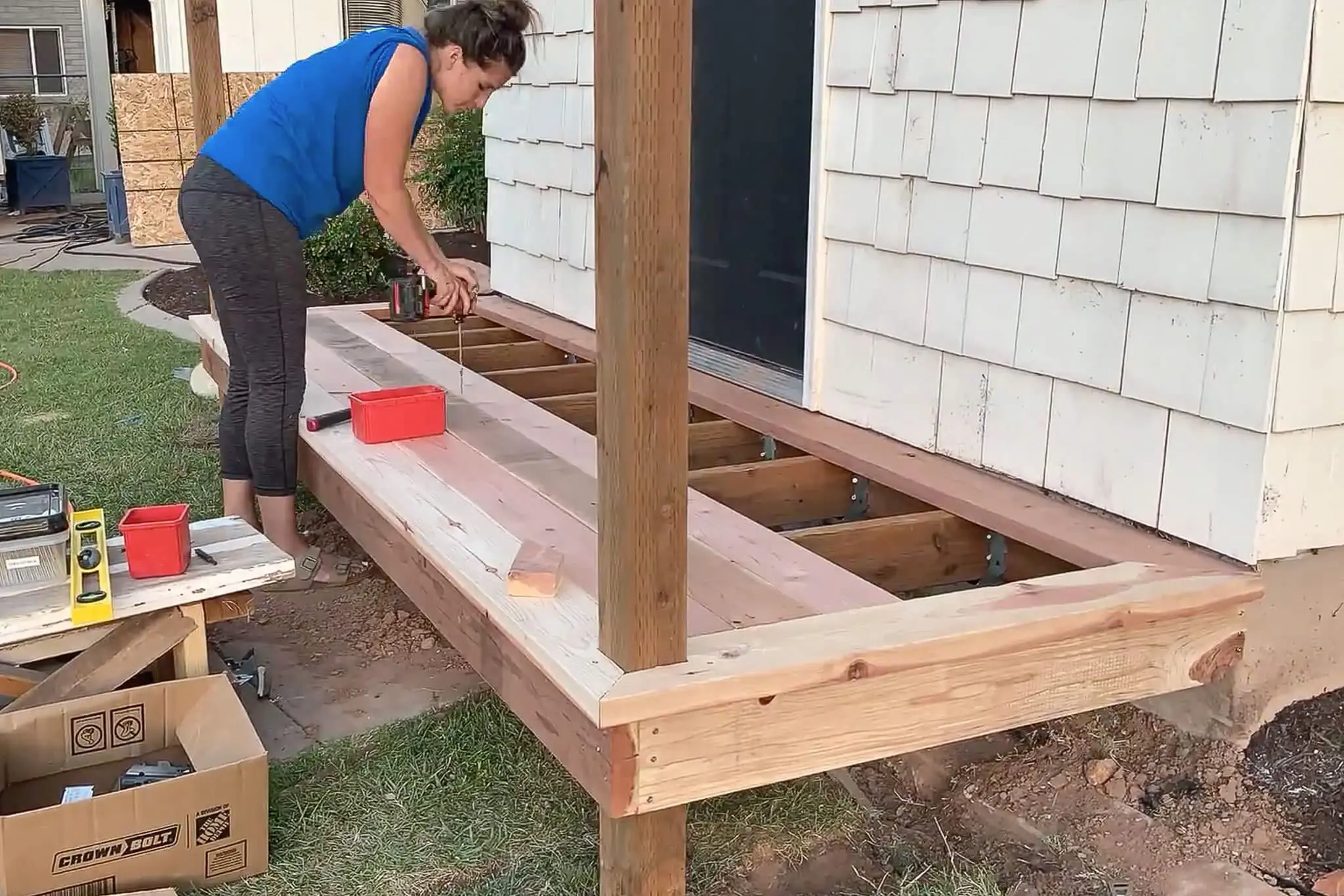 Woman drilling deck boards into place during a DIY small front porch project, securing each board over the exposed joists with tools nearby.