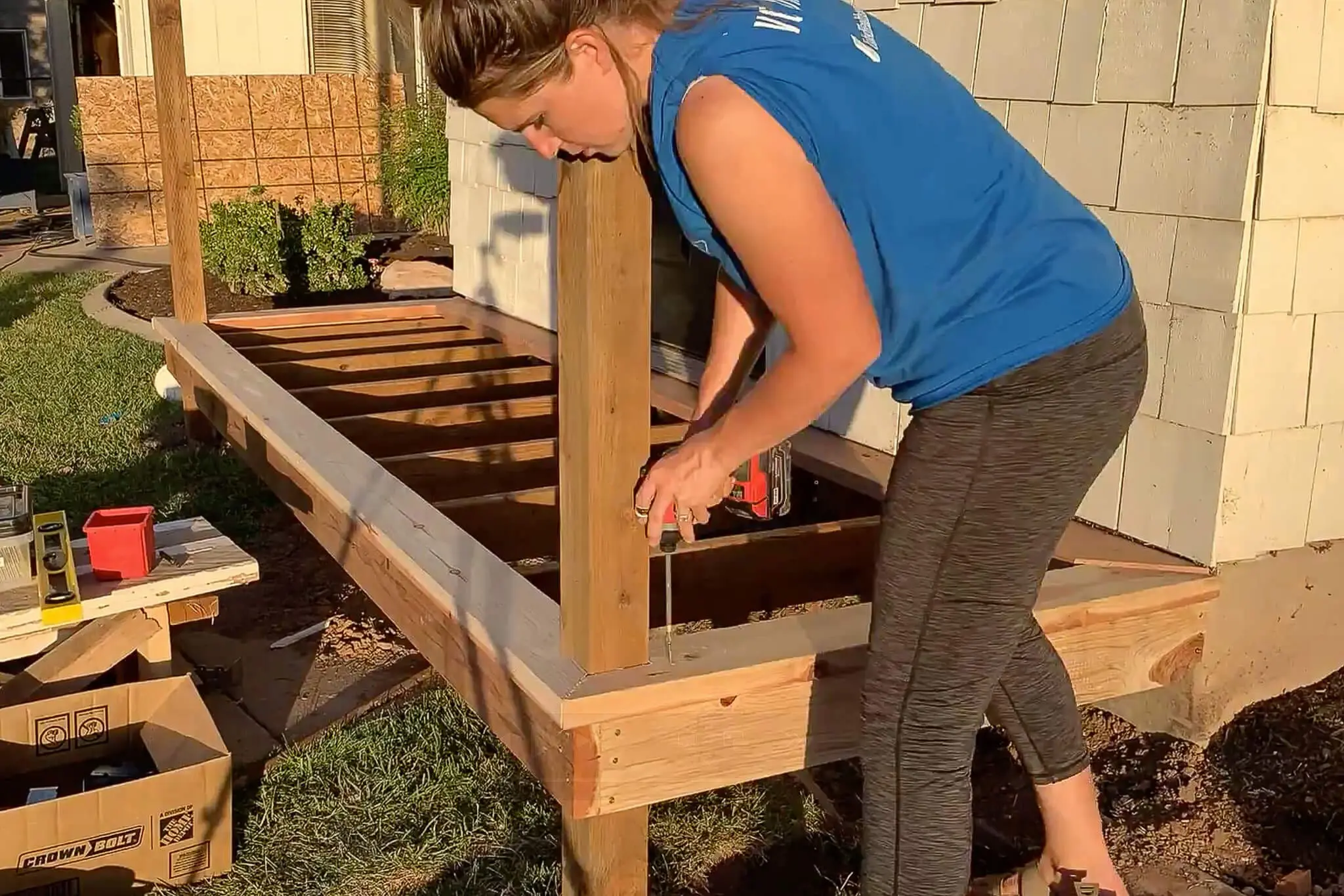 Woman using a drill to install trim boards on the frame of a porch, with exposed joists and porch posts during construction.