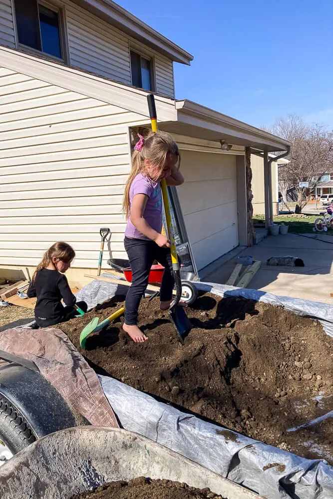 Young girl holding two small gardening shovels while smiling in a backyard garden setup, ready to help with planting.

