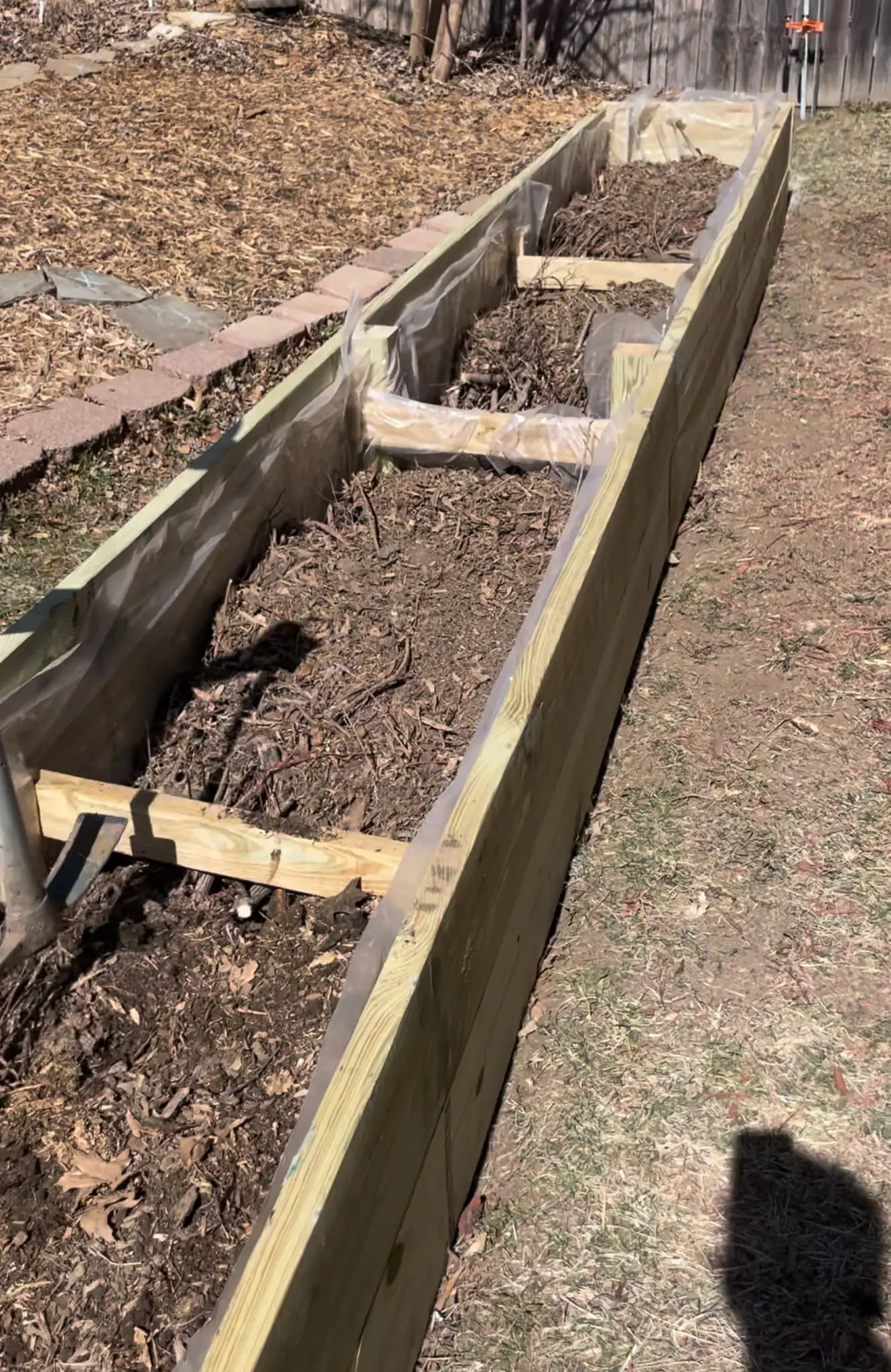 Backyard garden with tall wooden planter boxes filled with soil, sitting against a white house with bright green lawn in the foreground.