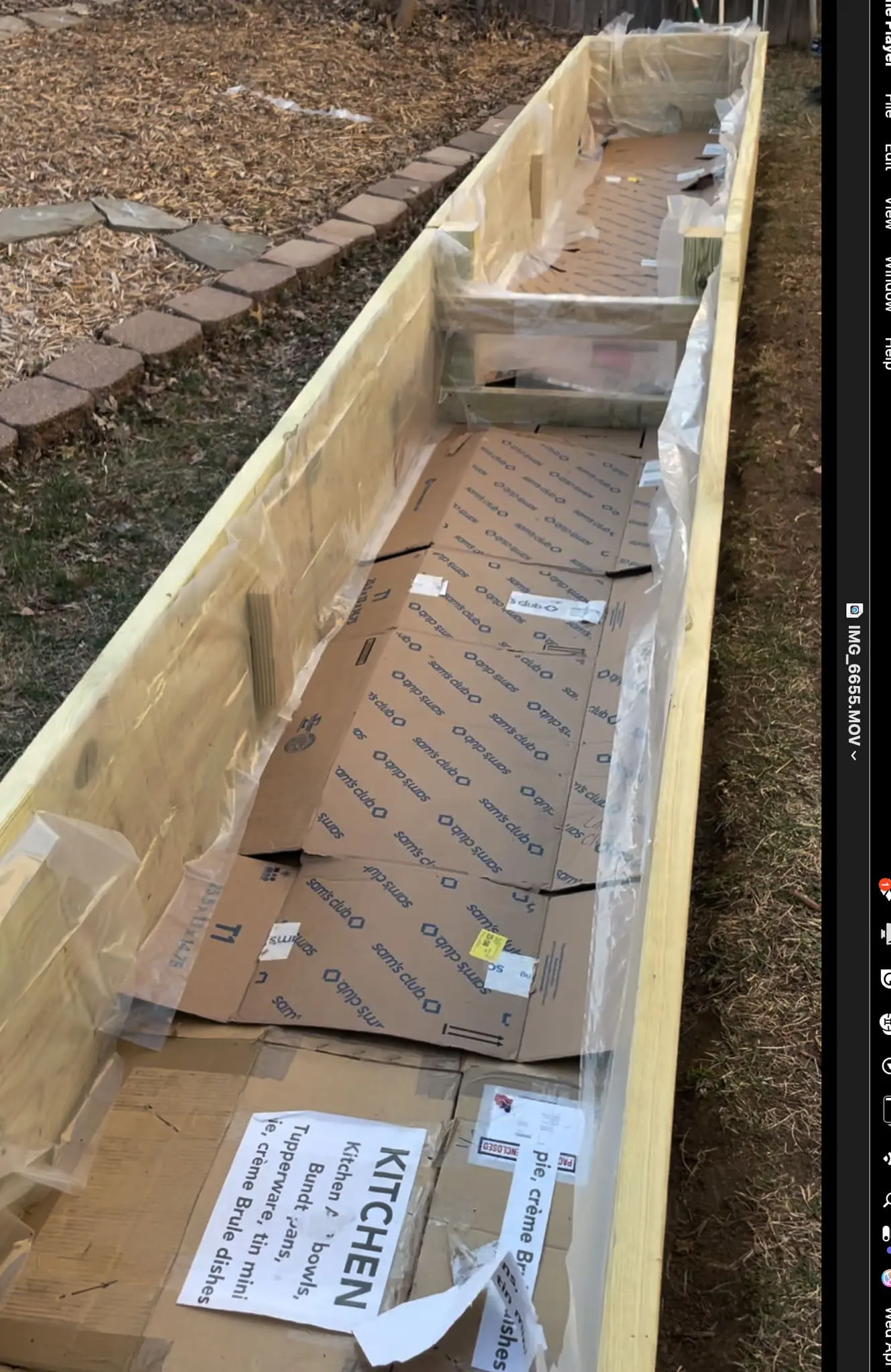 Raised garden bed filled with soil and layered with cardboard and wood debris, illustrating an efficient small-space gardening setup along the side of a house.