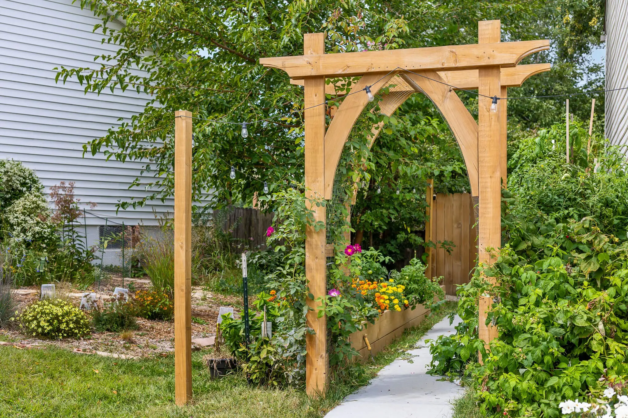 Arched wooden garden arbor built over a concrete path leading into a lush vegetable garden with raised planter boxes and colorful flowers