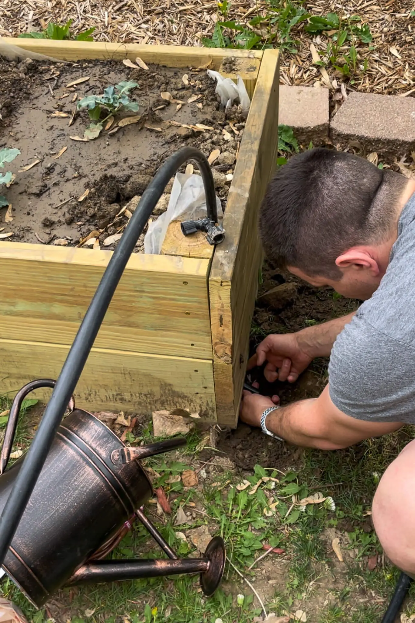 A person connecting a black drip irrigation line to a 90-degree corner fitting at the edge of a wooden raised garden bed.