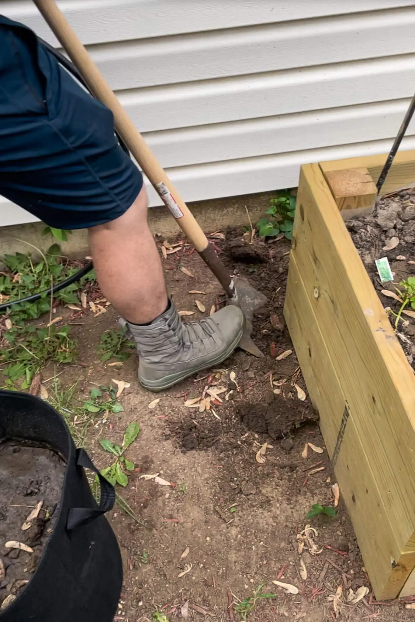 A person digging a narrow trench beside a wooden raised garden bed, preparing to install a drip irrigation system.