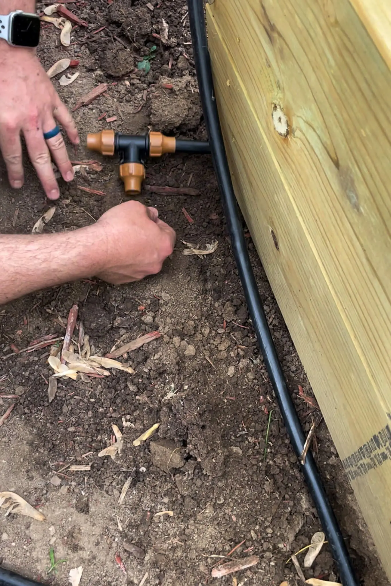 A person installing a T-fitting into a black drip irrigation hose beside a wooden raised garden bed.