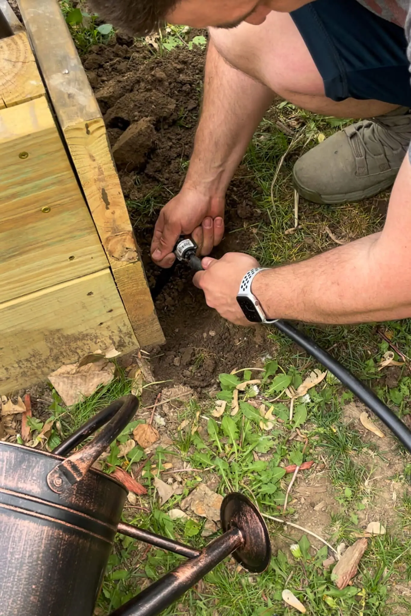  Close-up of hands securing a black drip irrigation line to a barbed connector at the edge of a wooden raised garden bed.