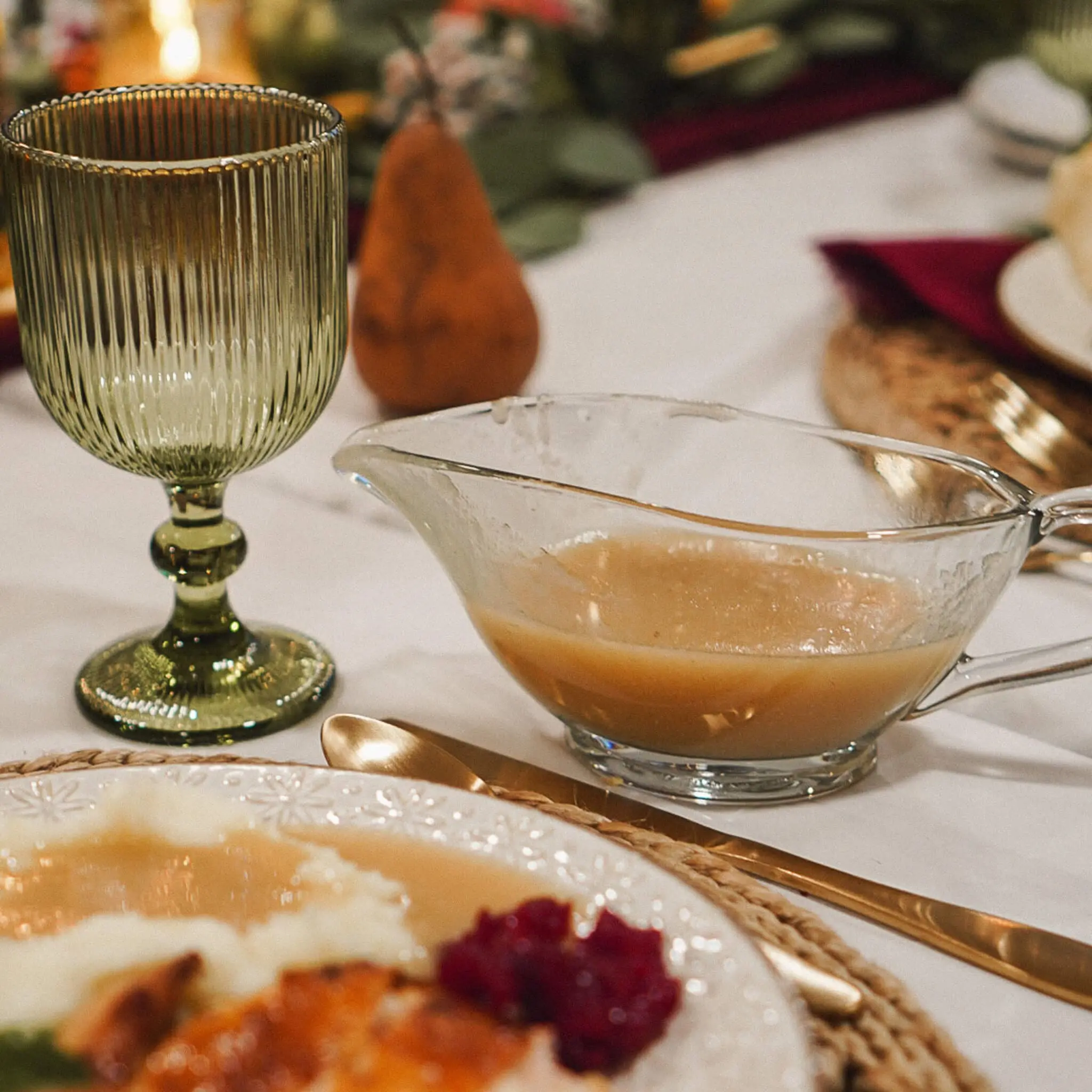 A glass gravy boat filled with rich turkey gravy on a festive holiday table, alongside a green ribbed glass goblet, a plate of turkey with cranberry sauce, and seasonal decor in the background.