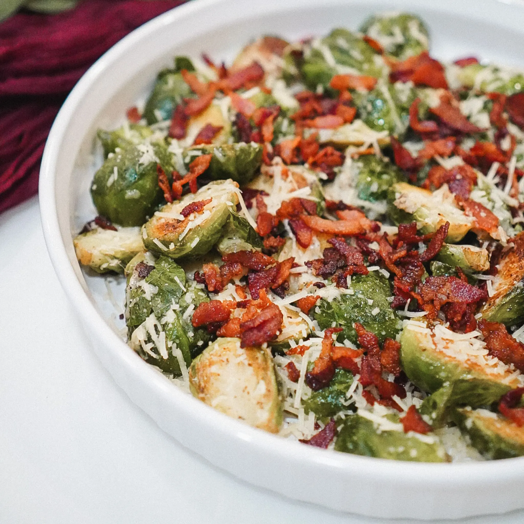 A white serving dish filled with halved Brussels sprouts sautéed to golden perfection, topped with crispy bacon pieces and shredded Parmesan cheese—an easy and flavorful vegetable side dish.