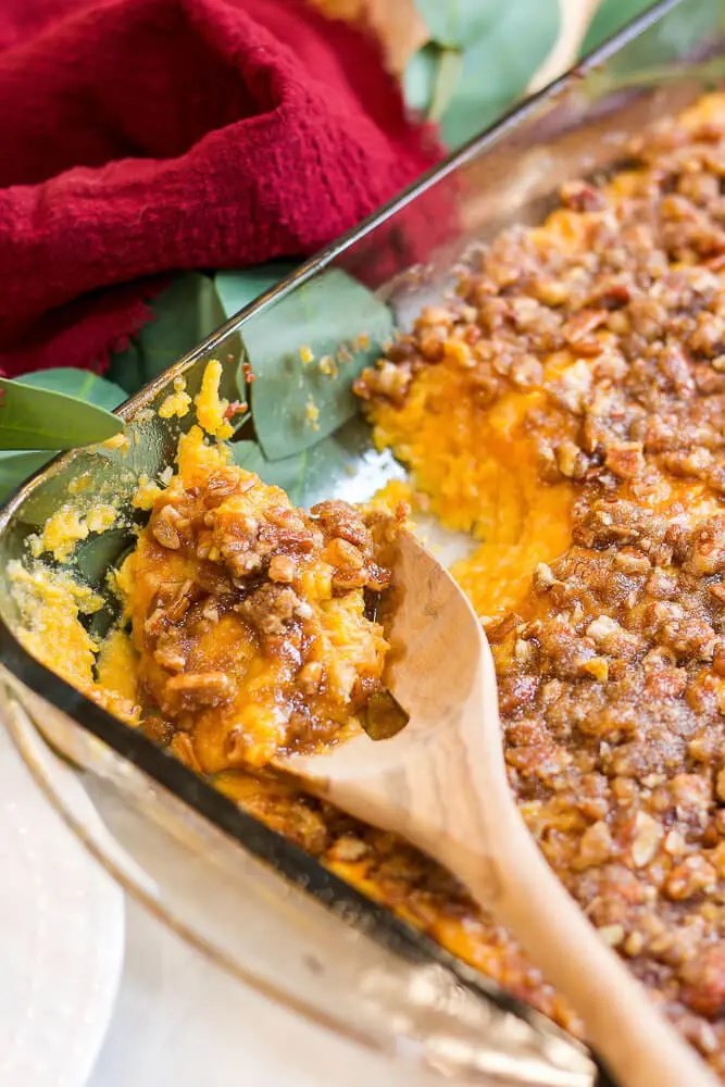 A spoon scooping out a serving of sweet potato casserole with a pecan crumble, the rich orange and brown colors contrasting with a red napkin in the background.