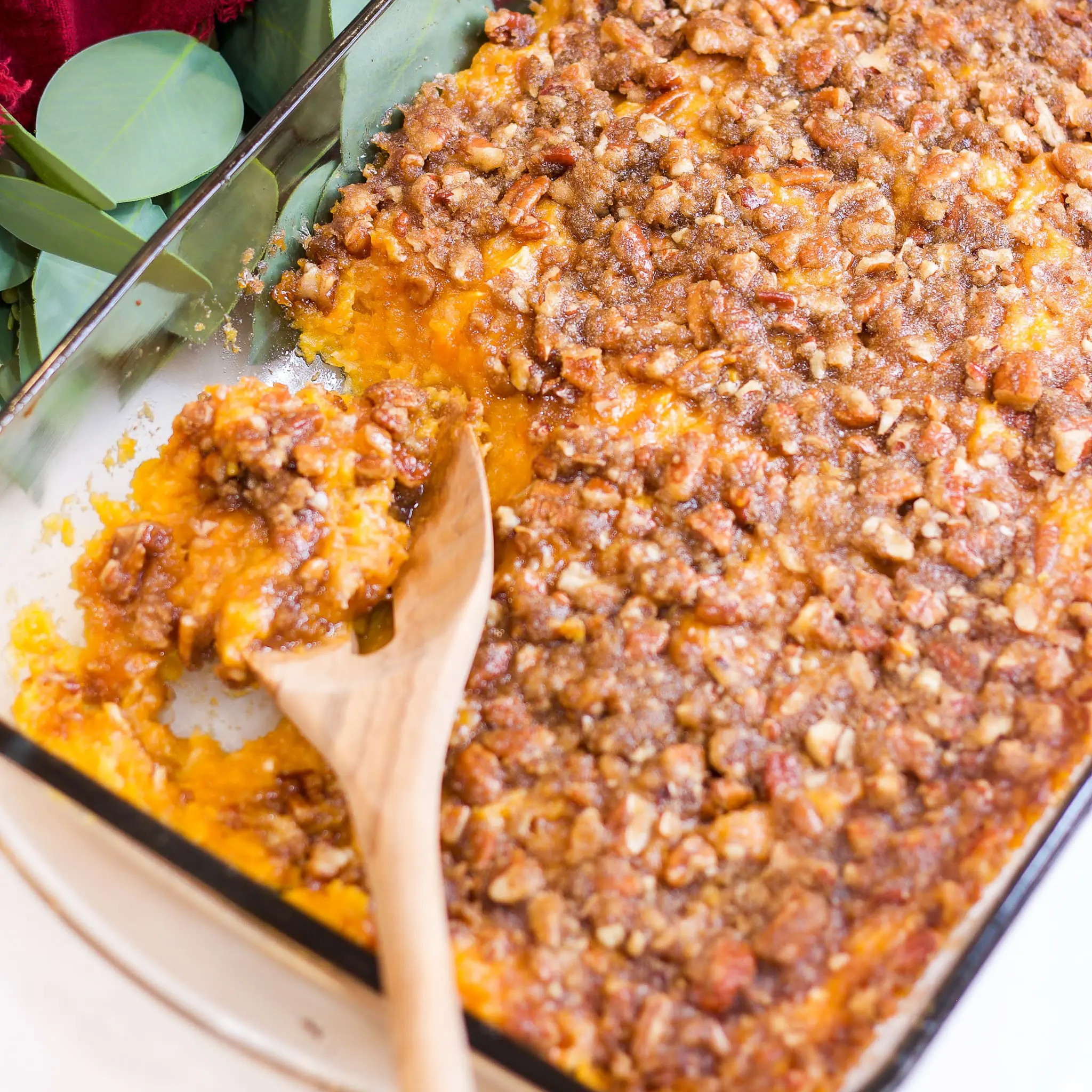 A glass baking dish filled with sweet potato casserole topped with a golden-brown pecan and brown sugar crumble, with a wooden spoon scooping out a serving—perfect for Thanksgiving or holiday meals.