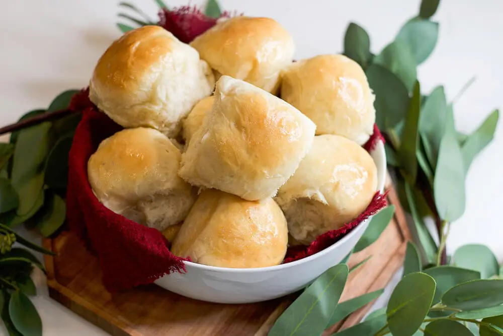 A bowl filled with soft, golden dinner rolls nestled in a red cloth, surrounded by green eucalyptus leaves for a beautiful presentation.