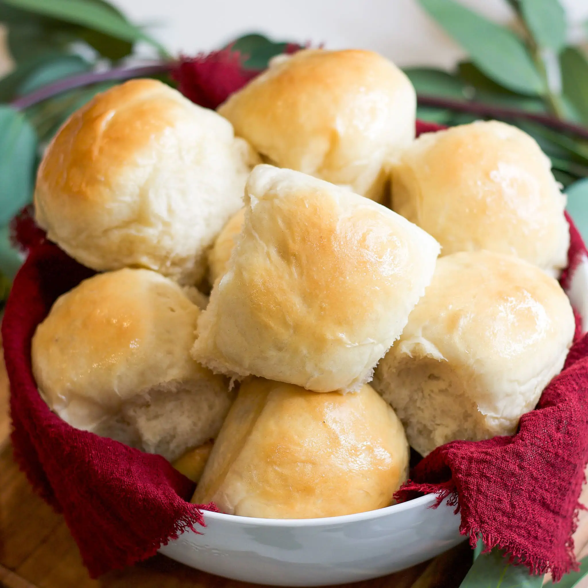 A bowl of golden, fluffy homemade dinner rolls nestled in a red cloth napkin, perfect for serving at holiday meals or family dinners.