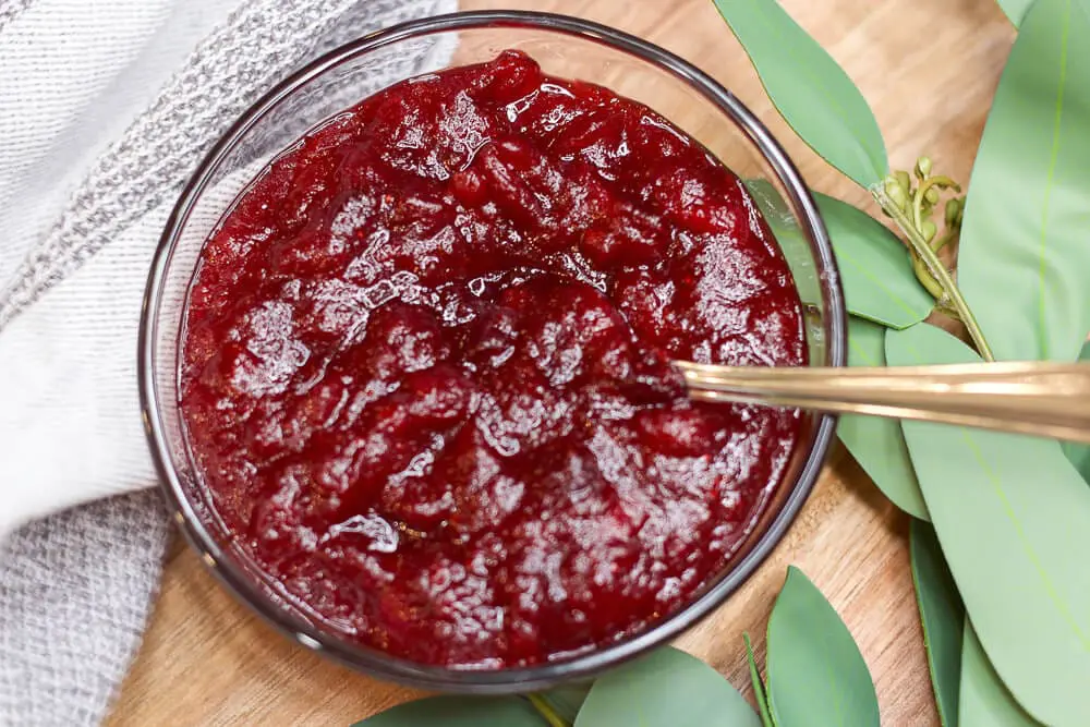 A glass bowl filled with thick homemade cranberry sauce, set against a wooden background with green leaves for decoration.