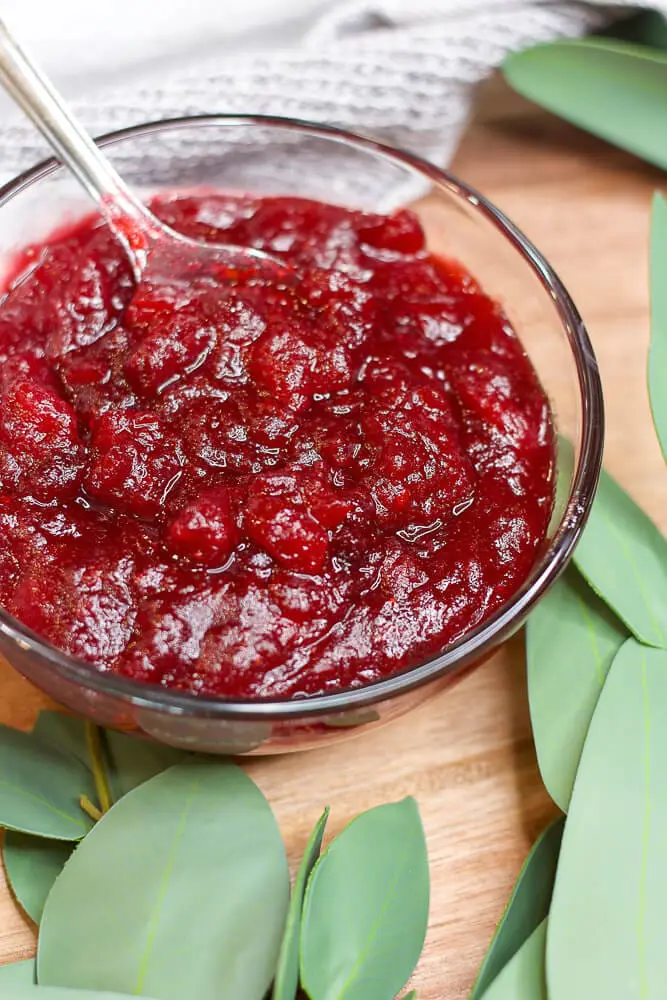 A glass bowl containing rich red cranberry sauce, with a spoon dipped inside, placed on a wooden surface with green leaves surrounding it.