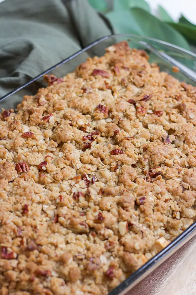 A close-up of golden-brown apple crisp with pecans baked in a glass dish, set on a wooden surface with a green cloth napkin in the background.