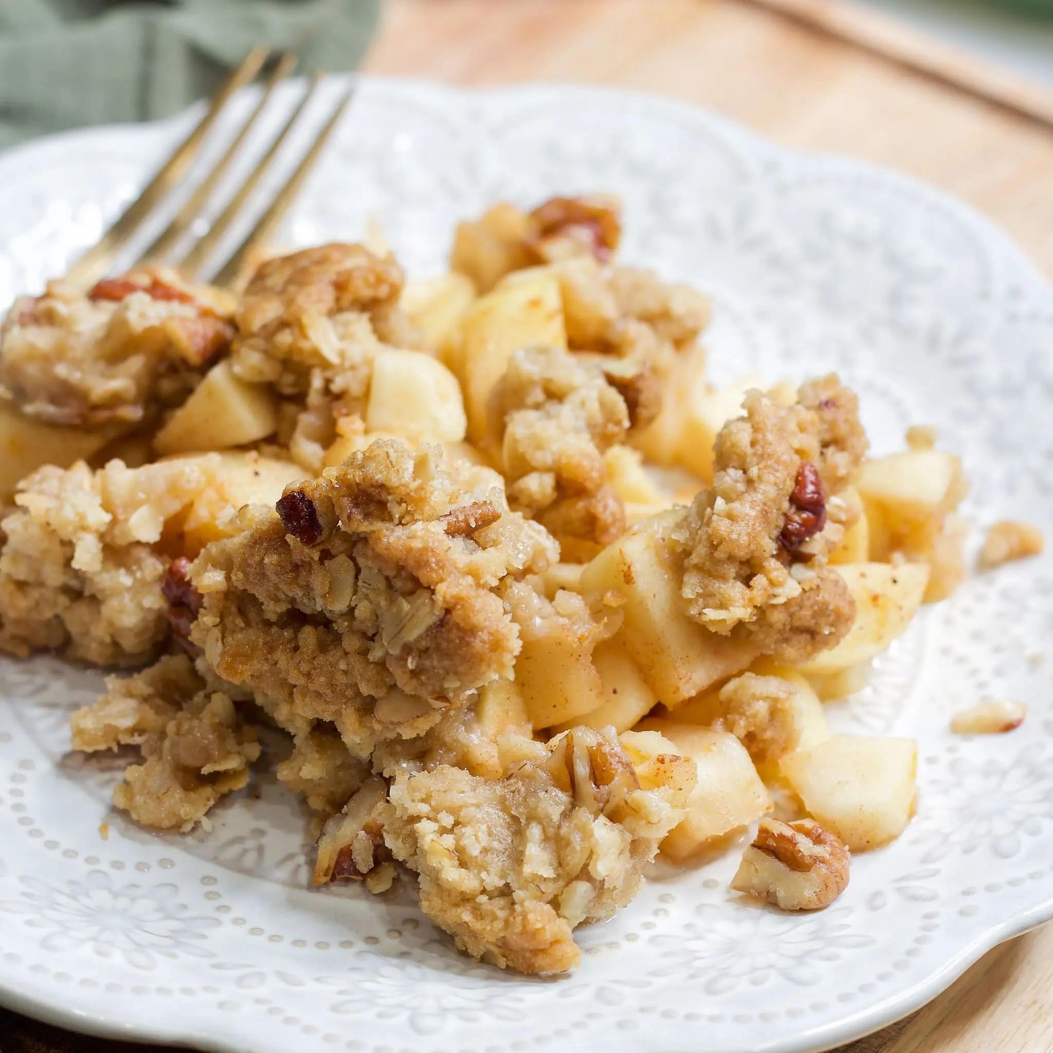 A serving of warm apple crisp with a buttery oat topping and chunks of baked apples, served on a white decorative plate—perfect cozy dessert inspired by Ina Garten.