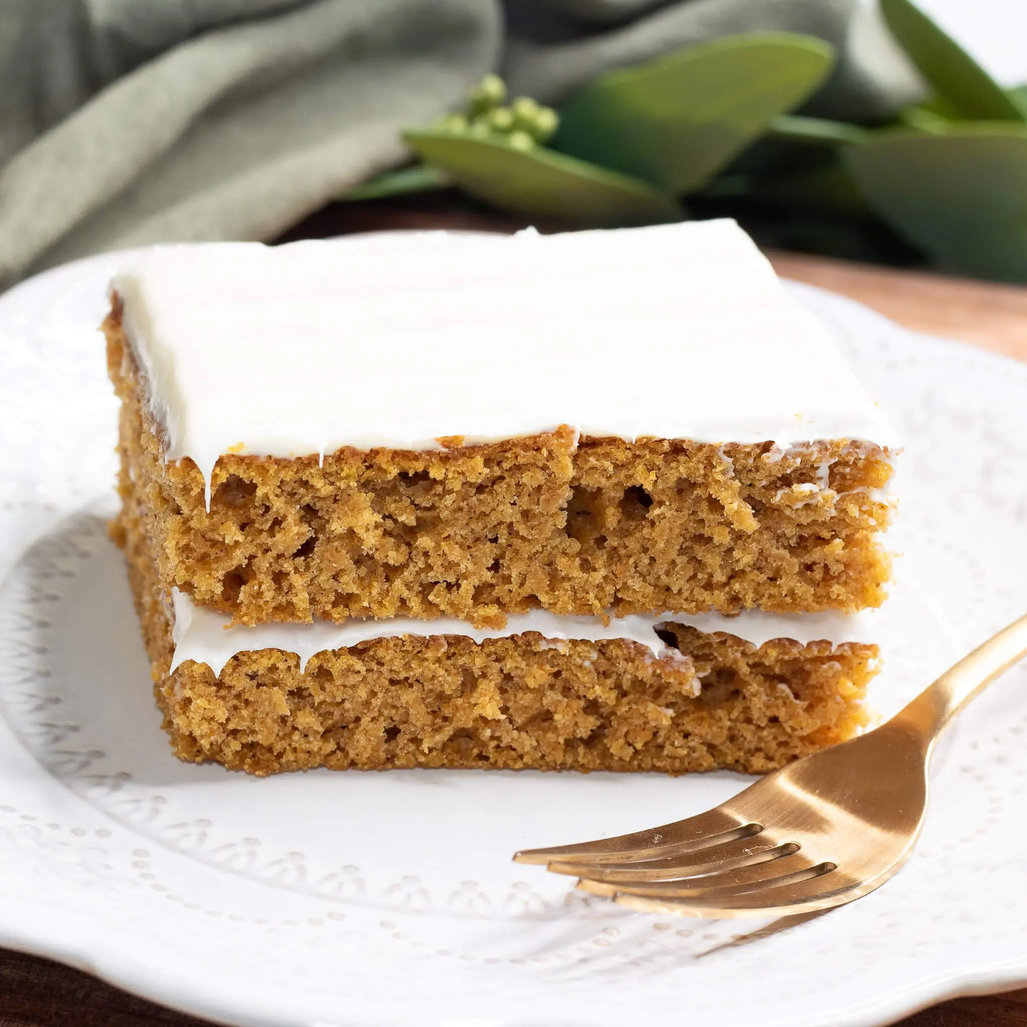 A moist slice of pumpkin cake with cream cheese frosting, layered and served on a white decorative plate with a gold fork—perfect for fall desserts or Thanksgiving celebrations.