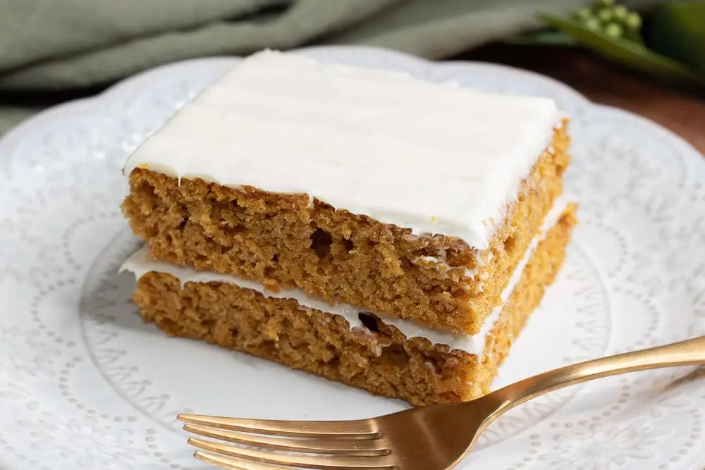 A square slice of pumpkin cake layered with cream cheese frosting on a white plate, accompanied by a gold fork.