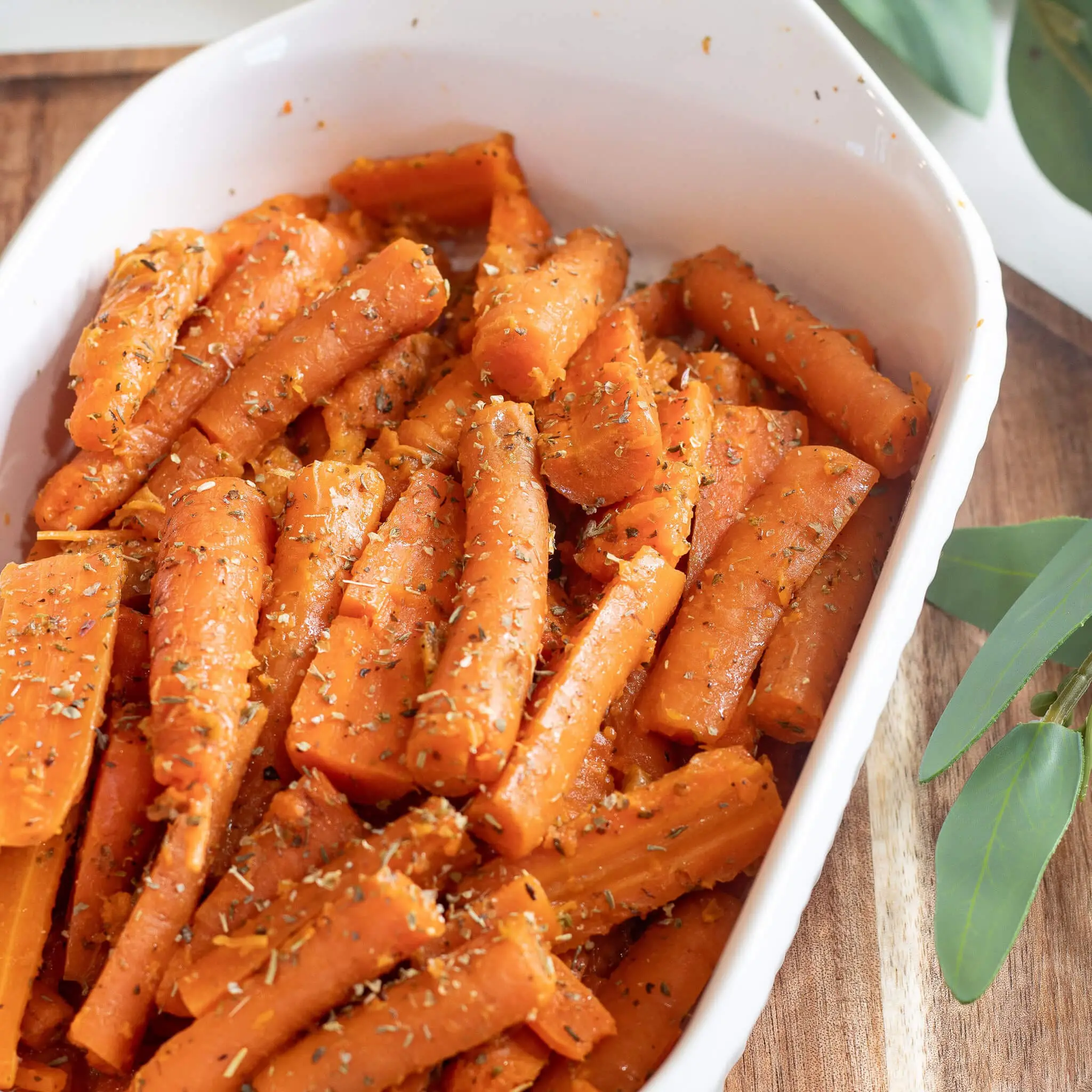 A white baking dish filled with tender steamed carrots, cut into rustic pieces and tossed with melted butter and Italian seasoning—served as a flavorful, simple vegetable side dish.
