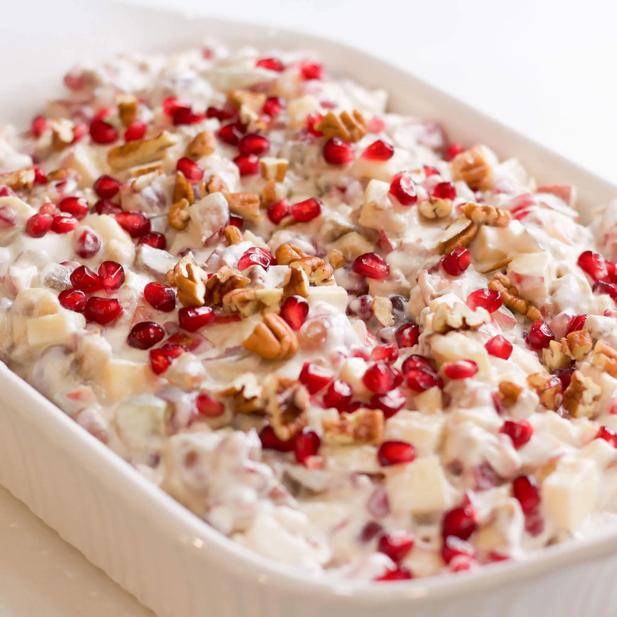 A white serving dish filled with creamy pomegranate salad, topped with bright red pomegranate seeds and chopped pecans—showcasing a festive mix of fruit and whipped cream, perfect for holiday gatherings.