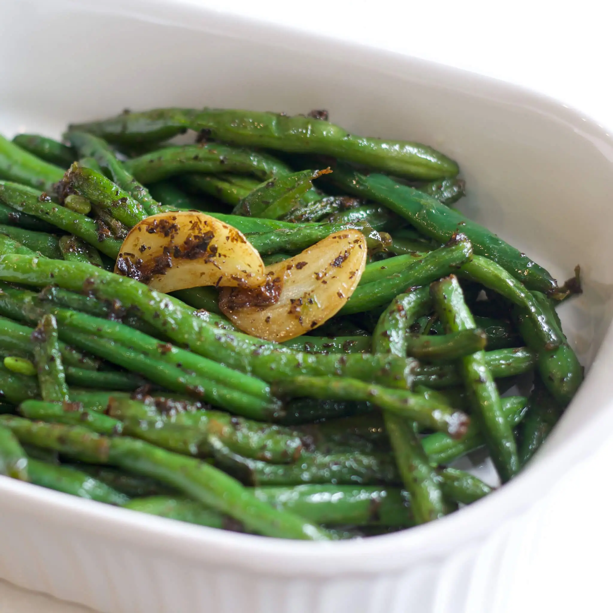 A white serving dish filled with sautéed fresh green beans, glistening with butter and olive oil, and topped with caramelized garlic slices and herbs—perfect for a flavorful holiday side dish.