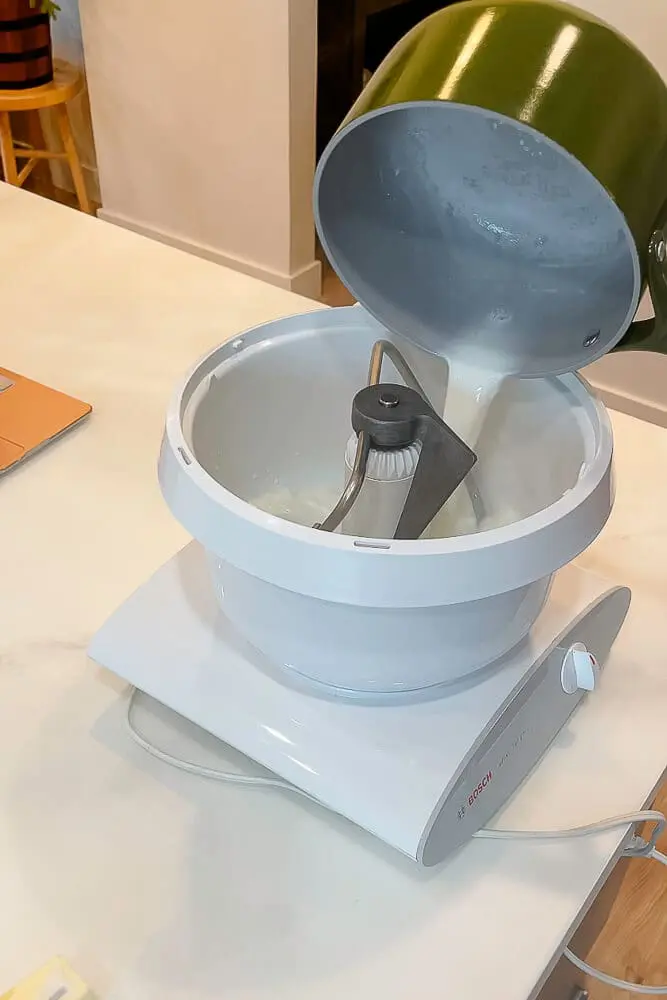 Warm milk being poured from a saucepan into a stand mixer, preparing dough for homemade dinner rolls.